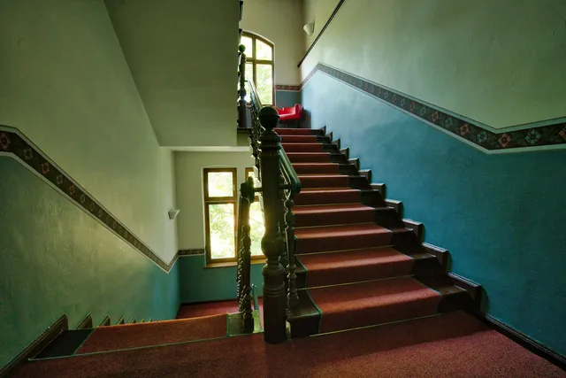 A worn stairwell in an older apartment block, green painted walls and a wooden handrail turning out of sight
